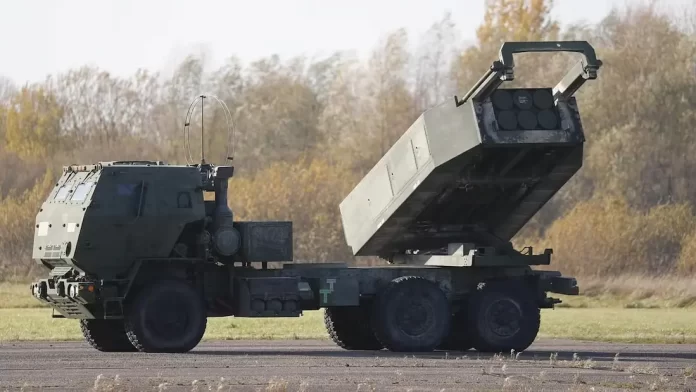 Military mobile launcher vehicle with a raised rocket/missile launcher on a wheeled chassis in an open field.