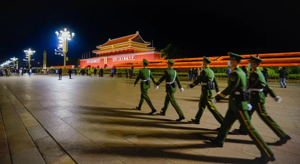 di notte una fila ordinata di soldati in uniforme verde marcia lungo la vasta piazza tiananmen illuminata con sullo sfondo la porta di tiananmen colorata di rosso e decorata da grandi cartelli luminosi