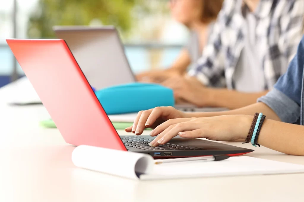 una persona seduta a una scrivania che digita su un laptop sottile di colore rosso con davanti un quaderno bianco arrotolato sul tavolo
