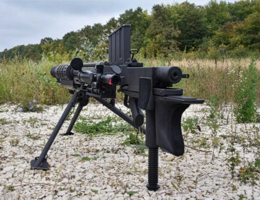 military machine gun mounted on a tripod in a grassy gravelly field with trees in the background