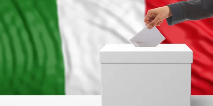 Una mano inserisce una scheda in un’urna bianca davanti a una grande bandiera italiana.