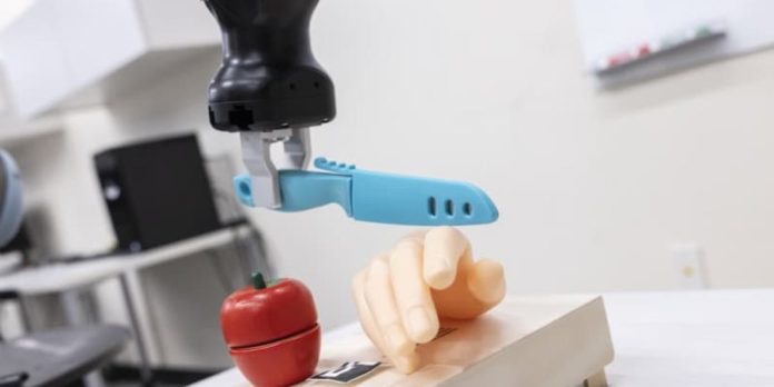 Braccio robotico che tiene un coltello giocattolo azzurro sopra una mano finta, accanto a una mela di legno su un tagliere