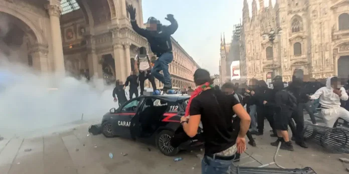 Manifestanti saltano su un'auto dei carabinieri in piazza del Duomo a Milano, tra fumogeni e transenne rovesciate