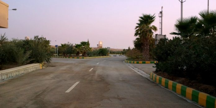 Strada deserta che conduce a un valico di frontiera tra Iraq e Iran, con palme e posto di controllo sullo sfondo
